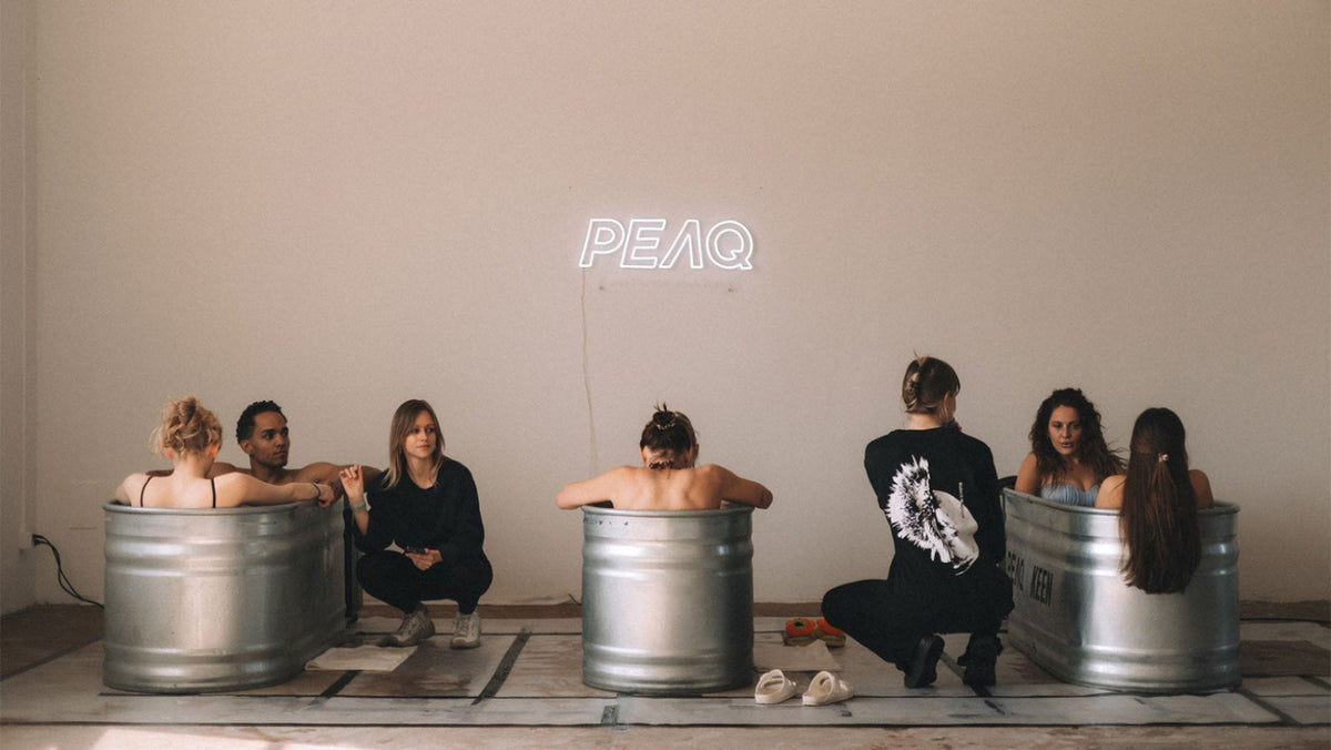 A group of people relax in large galvanized steel tubs arranged in a room, with a glowing white neon sign reading 'PEAQ' mounted on the wall behind them.